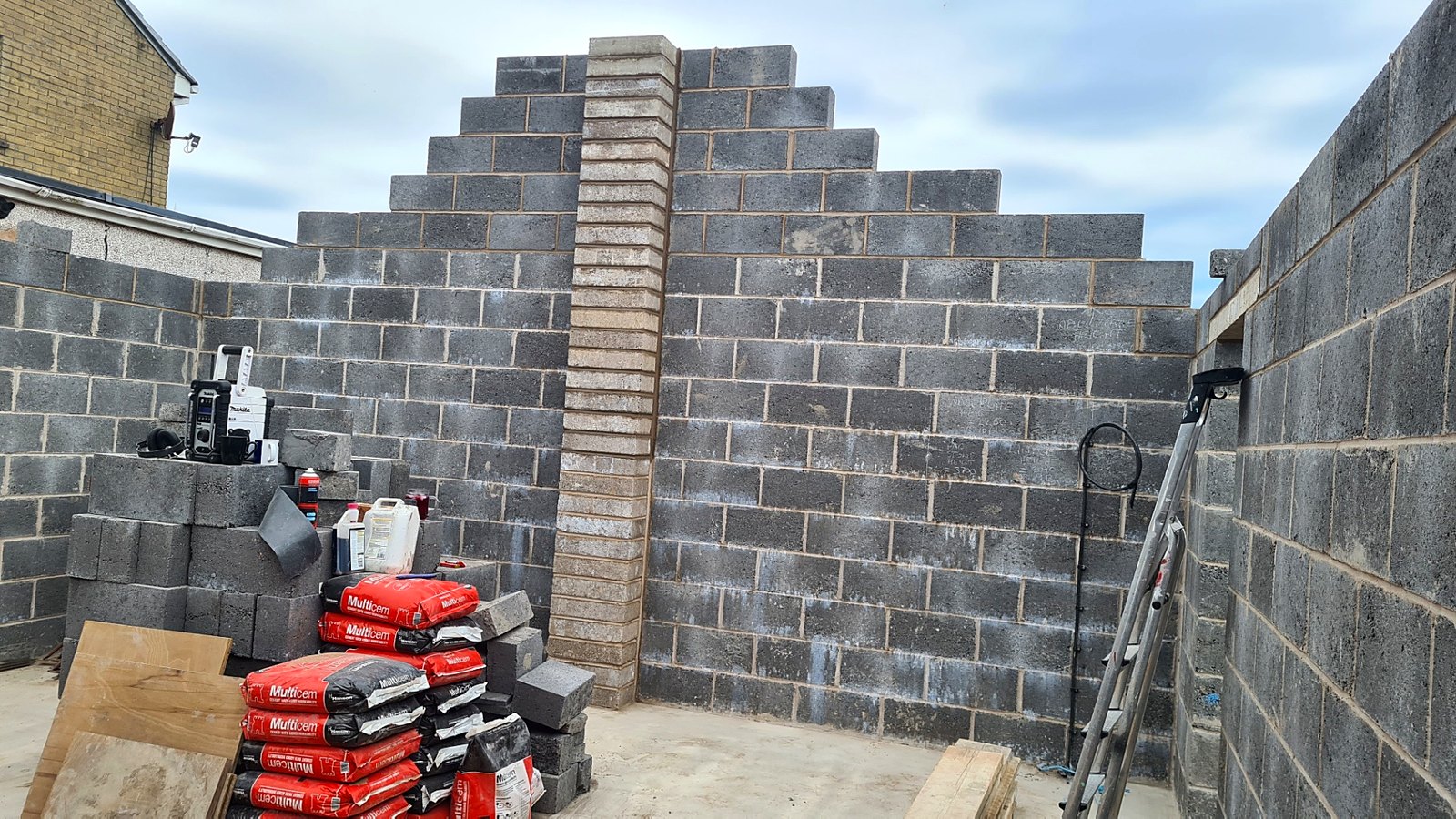 Blockwork gable wall with a brick pier under construction, cement bags stacked in the foreground