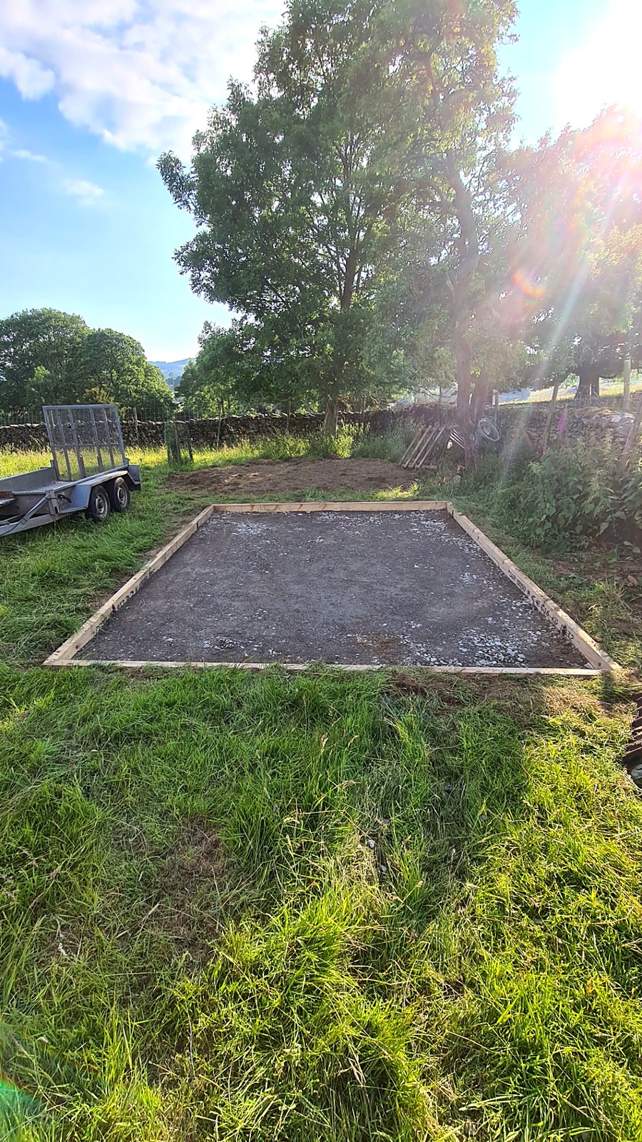 Compacted stone base within timber shuttering prepared in a rural grassed area beside a plant trailer