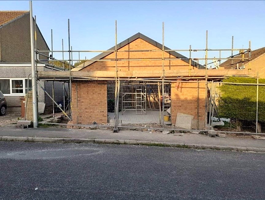 Brickwork shell of a single-storey residential building with a timber gable frame and scaffolding around it